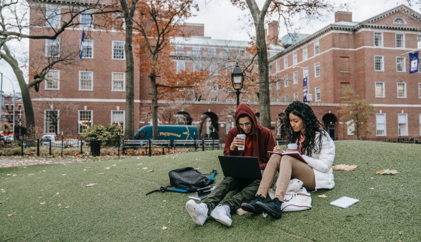 Two students sit on grass outside of a red brick university building