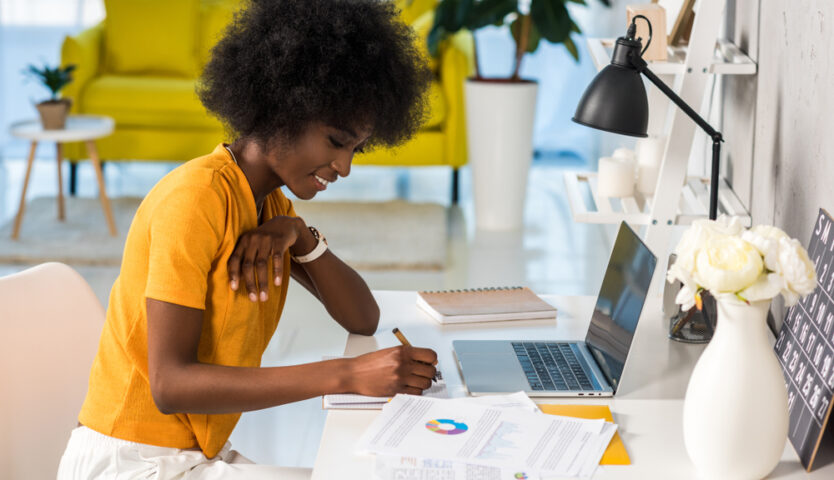 black woman in yellow shirt sitting at desk with laptop doing tax paperwork