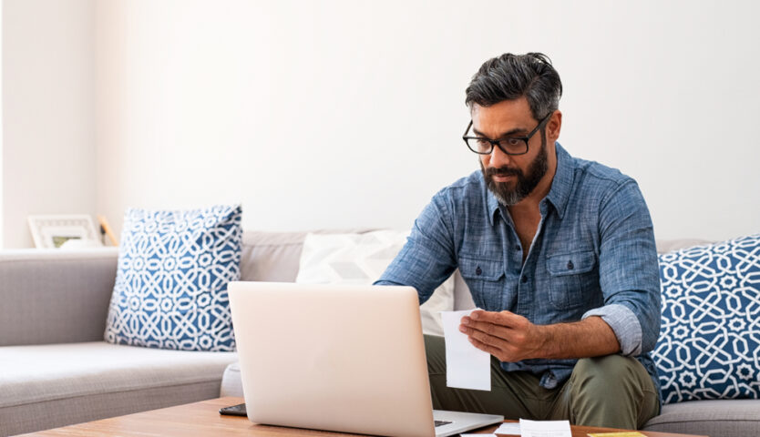 man with beard and glasses wearing denim shirt sitting a grey couch doing his taxes on his laptop