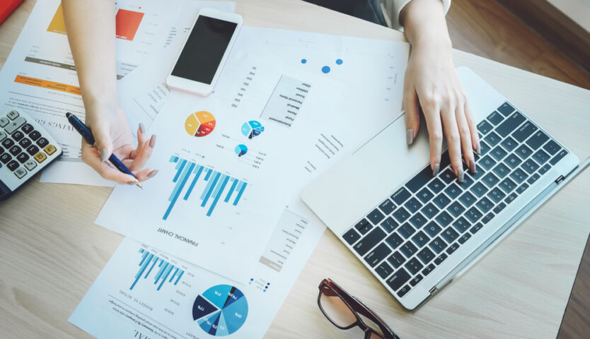 person with long taupe nails using a laptop while looking at paperwork with graphs and charts.