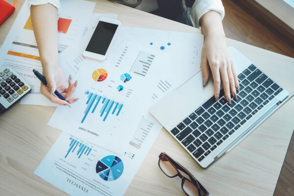 person with long taupe nails using a laptop while looking at paperwork with graphs and charts.