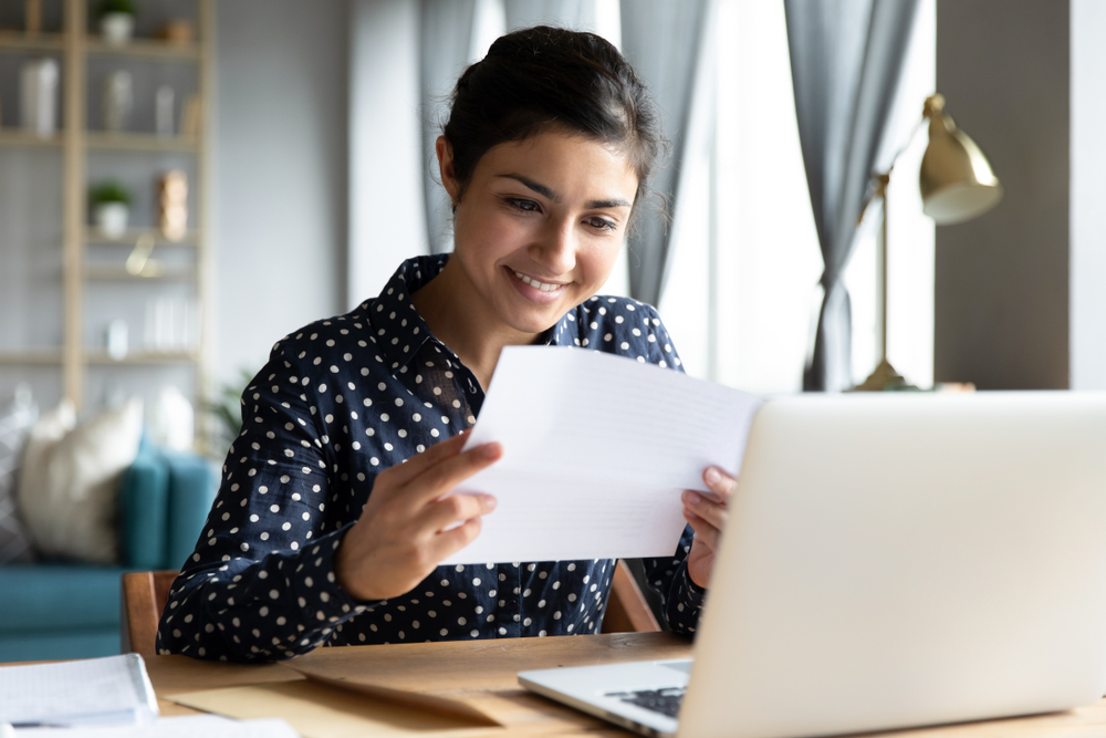 Smiling woman sitting in front of laptop looking at piece of paper.