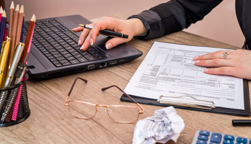 overhead photo of hands working on a laptop with eye glasses on desk and pencil holder on desk. crumbled paper is nearby.