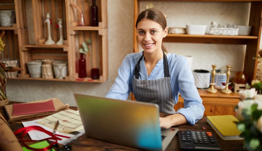 female small business owner using laptop and looking at camera in shop