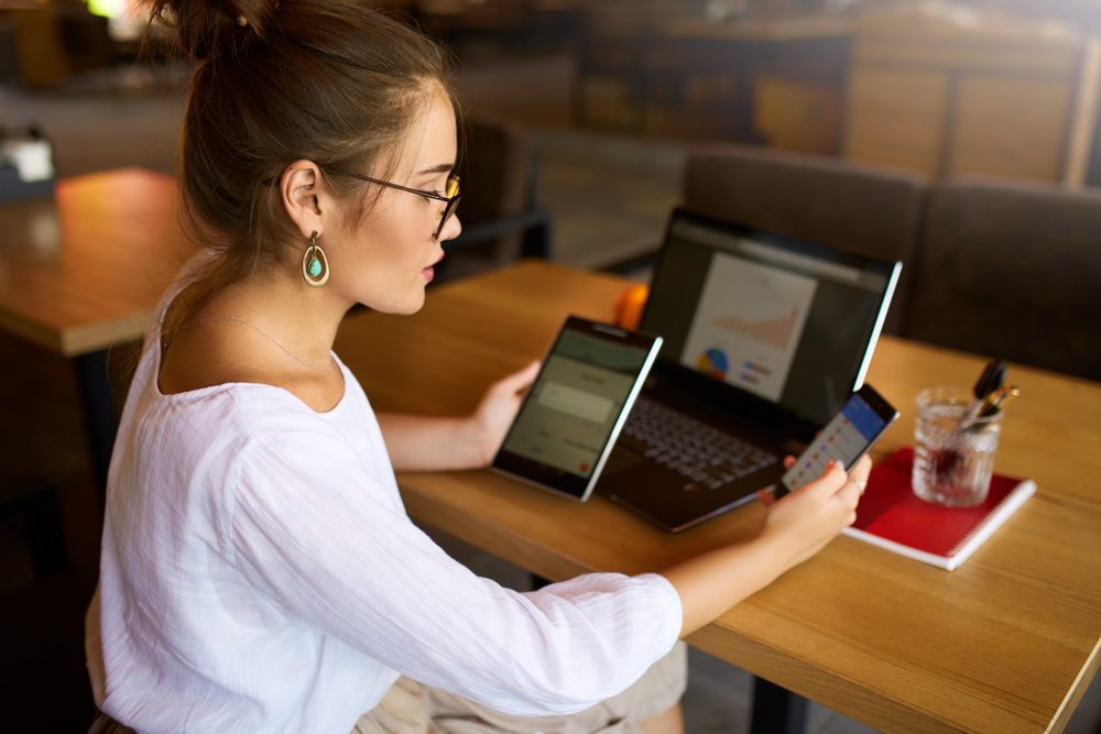 Freelancer businesswoman has tablet and cellphone in hands and laptop on table with charts on screen.